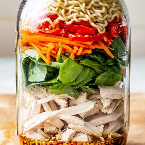 Clear glass mason jars stacked with colorful layers of noodles, fresh vegetables, and diced chicken in a savory sauce.