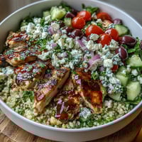 Freshly grilled lemon-oregano chicken strips and crumbled feta top a vibrant bowl of Mediterranean Pearl Couscous Chicken Bowls with tomatoes and cucumbers.